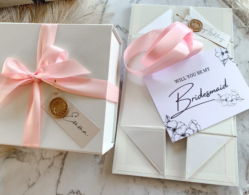 May include: A white gift box with a pink ribbon and a tag that says "Emma". The box is next to a white box with a pink ribbon and a card that says "Will you be my Bridesmaid?" with a floral design.