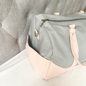 May include: A grey canvas duffel bag with a light pink leather base and accents. The bag has a zipped top, a side pocket, and light pink handles. The bag is designed for travel or everyday use.