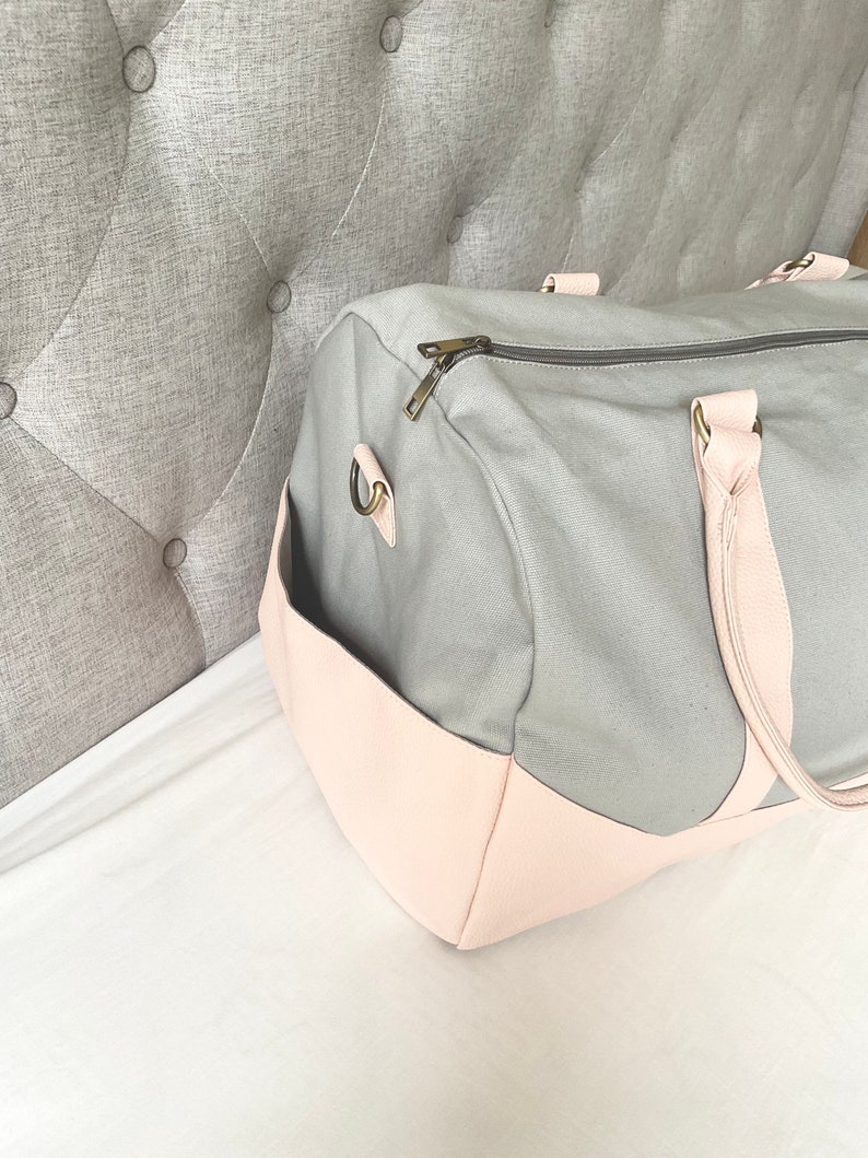 May include: A grey canvas duffel bag with a light pink leather base and accents. The bag has a zipped top, a side pocket, and light pink handles. The bag is designed for travel or everyday use.
