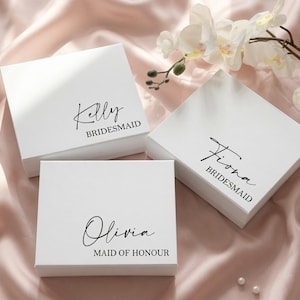 May include: Three square white gift boxes with black text. The top left box reads Kelly BRIDESMAID, the top right box reads Fiona BRIDESMAID, and the bottom box reads Olivia MAID OF HONOUR in a combination of script and block lettering.