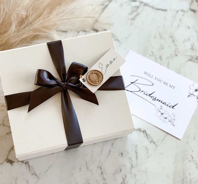 May include: A white gift box with a black satin ribbon and a gold wax seal. The box has a tag attached that says "Emma". A white card with black text says "Will you be my Bridesmaid".
