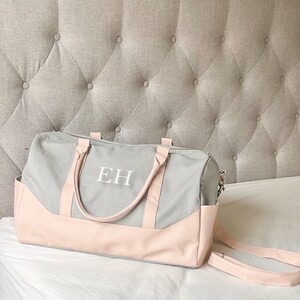 May include: A grey and pink duffel bag, featuring the white initials "EH". The bag has a grey main body with pink detailing on the base and handles. A detachable pink shoulder strap is also included.
