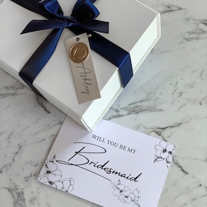 May include: A white gift box with a blue ribbon and a tag that says "Ashley". The box is sitting on a white marble surface next to a white card with black text that says "Will you be my Bridesmaid" and a floral design.