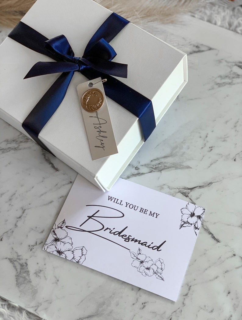 May include: A white gift box with a blue ribbon and a tag that says "Ashley". The box is sitting on a white marble surface next to a white card with black text that says "Will you be my Bridesmaid" and a floral design.