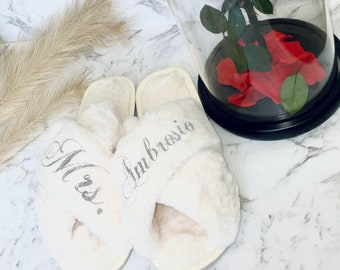 May include: A pair of cream-colored, fluffy cross-band slippers. The left slipper has the word "Mrs." embroidered in silver, and the right slipper has the name "Ambrosio" embroidered in silver. A preserved red rose is in a glass dome in the background.