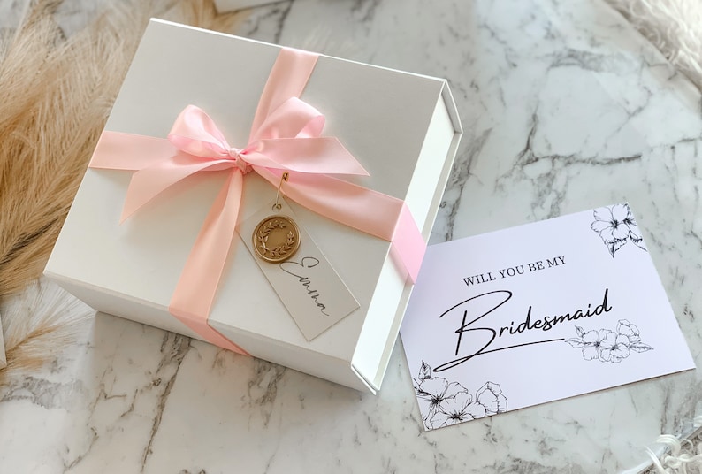 May include: A white gift box with a pink ribbon and a tag that says "Christina". The box is sitting on a marble surface next to a white card with the text "Will you be my Bridesmaid" and a floral design.