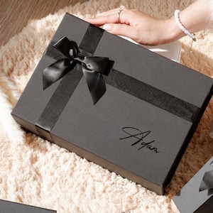 May include: A black gift box with a black satin ribbon and bow. The box has the name "Adam" written on it in a cursive font. The box is on a cream-colored, fluffy surface.