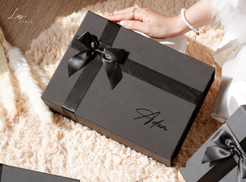 May include: A black gift box with a black satin ribbon and bow. The box has the name "Adam" written on it in a cursive font. The box is on a cream-colored, fluffy surface.