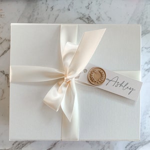 May include: A white gift box with a satin ribbon tied in a bow. The box has a tag with a gold wax seal and the name "Ashley" written on it.