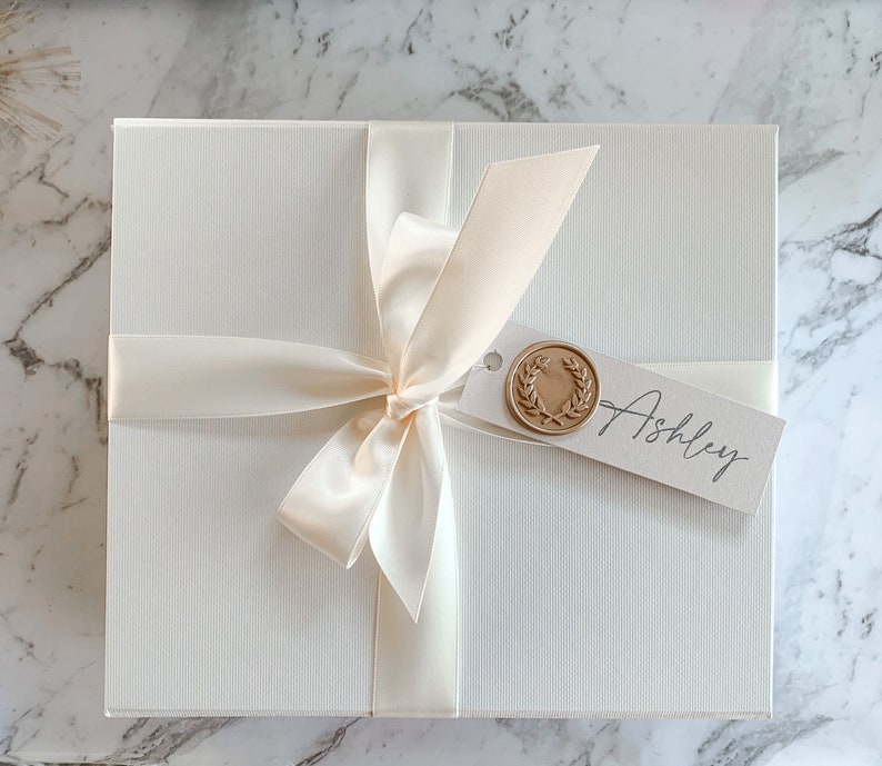 May include: A white gift box with a satin ribbon tied in a bow. The box has a tag with a gold wax seal and the name "Ashley" written on it.