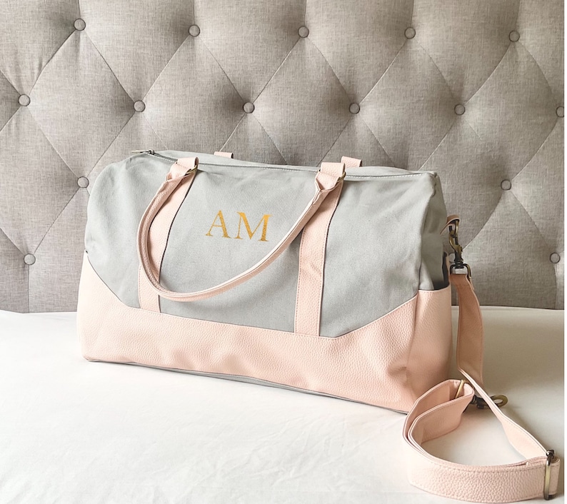 May include: A grey and pink duffel bag with the gold letters "AM" on the front. The bag has a light grey canvas body with pink faux leather accents on the bottom and handles. A detachable shoulder strap is also included.