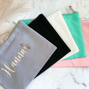 May include: A collection of canvas zipper pouches in various colors: gray with gold text, black, white, mint green, and pink. The gray pouch is personalized with the name "Hannah" in gold script. Each pouch has a gold zipper.