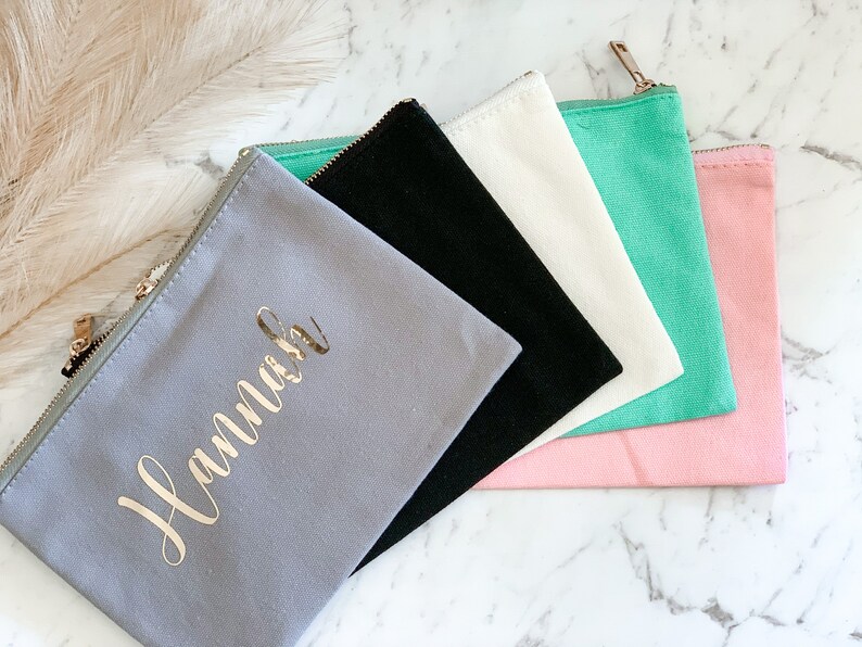 May include: A collection of canvas zipper pouches in various colors: gray with gold text, black, white, mint green, and pink. The gray pouch is personalized with the name "Hannah" in gold script. Each pouch has a gold zipper.