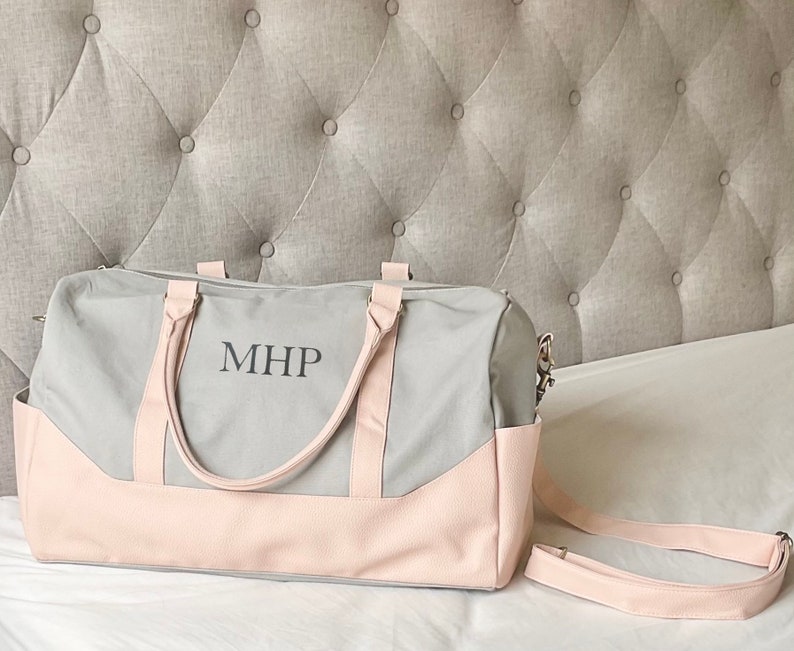 May include: A grey and pink duffel bag with the initials "MHP" printed on the front. The bag has a light pink base and handles, with a matching shoulder strap. The bag is on a white surface.