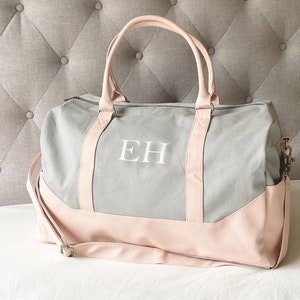 May include: A grey and pink duffel bag featuring the white initials "EH". The bag has pink handles and a shoulder strap. The base and accents are pink, with a grey main body. Ideal for travel or everyday use, it's a stylish accessory.
