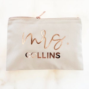 May include: A light gray zippered pouch with the words "mrs. COLLINS" in rose gold metallic lettering. The zipper and pull are also rose gold. The pouch is rectangular and appears to be made of canvas or a similar fabric.