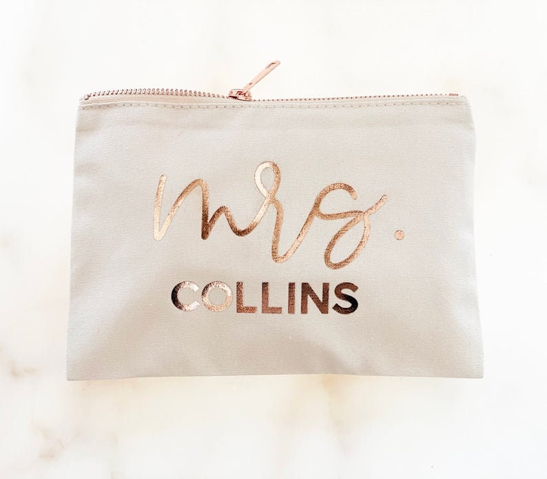 May include: A light gray zippered pouch with the words "mrs. COLLINS" in rose gold metallic lettering. The zipper and pull are also rose gold. The pouch is rectangular and appears to be made of canvas or a similar fabric.