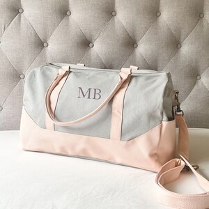 May include: A grey and pink duffel bag with the initials "MB" printed on the front. The bag features a light grey canvas body with pink faux leather accents on the bottom and straps. It includes a detachable shoulder strap.