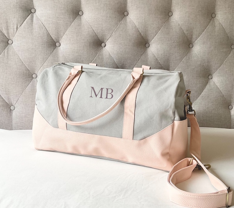 May include: A grey and pink duffel bag with the initials "MB" printed on the front. The bag features a light grey canvas body with pink faux leather accents on the bottom and straps. It includes a detachable shoulder strap.