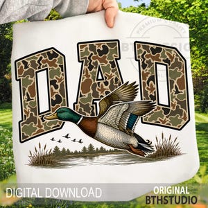 May include: White graphic with the word "DAD" in a camouflage pattern and black outline. A detailed illustration of a flying mallard duck is below the text, with a lake and reeds. The words "DIGITAL DOWNLOAD" and "ORIGINAL BTHSTUDIO" are also visible.