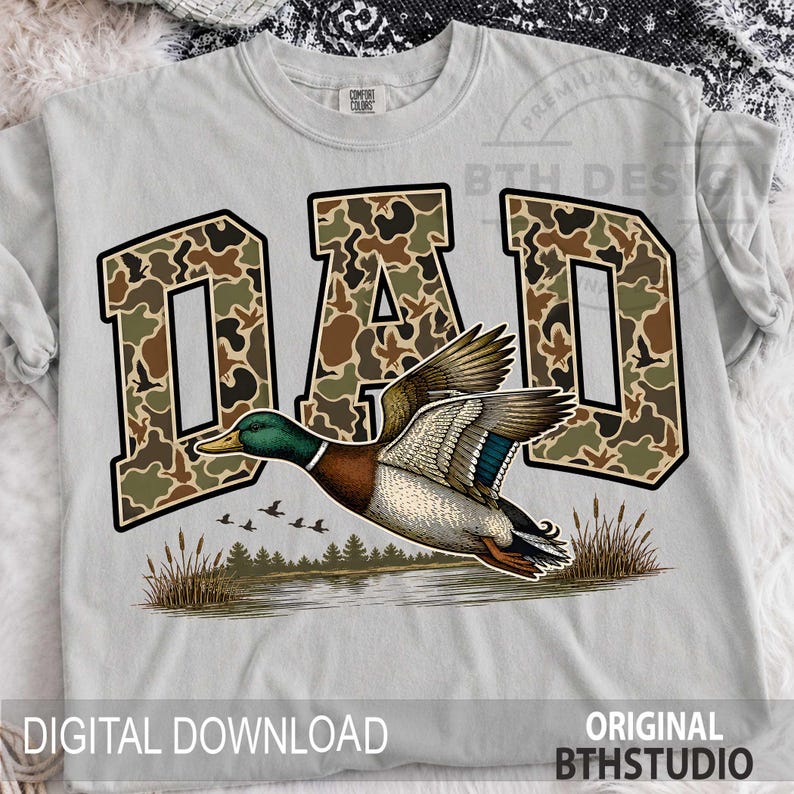 May include: A light grey t-shirt with the word "DAD" in a camouflage design and a black border. Below is a detailed illustration of a flying duck over a lake. The text "DIGITAL DOWNLOAD" and "ORIGINAL BTHSTUDIO" are at the bottom of the shirt.