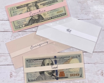 May include: Clear plastic envelopes with cash inside, labeled with categories: Cosmetics, HOA, Emergency, and Travel. The envelopes are designed for a personal financial planner.