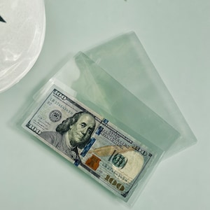 May include: A clear plastic sleeve with a $100 US bill inside. The bill features a portrait of Benjamin Franklin and the words "ONE HUNDRED DOLLARS" and "THE UNITED STATES OF AMERICA".