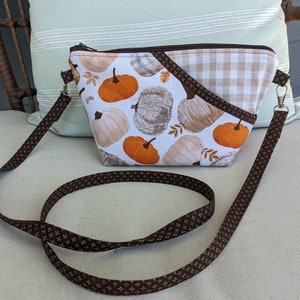 May include: A brown and white patterned crossbody bag with a removable strap. The bag features a white background with brown pumpkins and leaves. The flap of the bag is a brown and white gingham pattern.