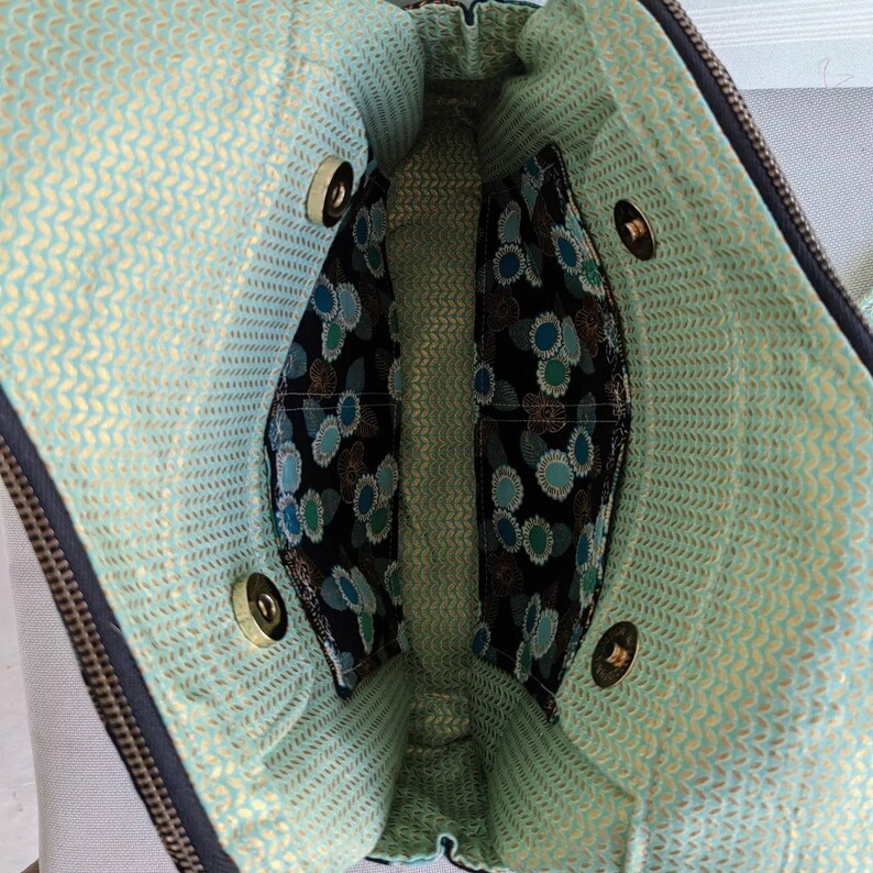 May include: The inside of a black bag with a green and gold patterned lining. The bag has two compartments and a zip closure. There are two gold snaps on each side of the bag.