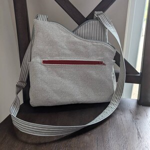 May include: A grey and white striped crossbody bag with a red zipper. The bag is made of a soft, textured fabric.