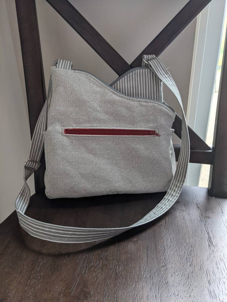 May include: A grey and white striped crossbody bag with a red zipper. The bag is made of a soft, textured fabric.