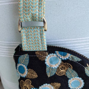 May include: Close-up of a black fabric bag with a gold and teal floral pattern. The bag has a teal and gold patterned strap with a brass D-ring.
