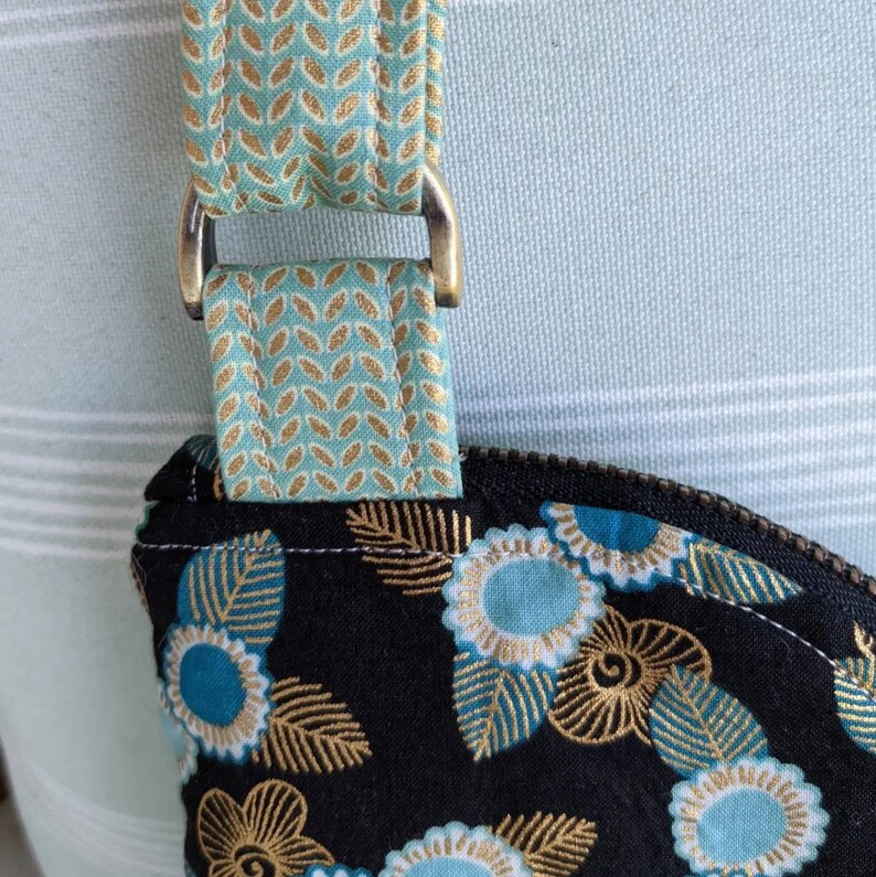 May include: Close-up of a black fabric bag with a gold and teal floral pattern. The bag has a teal and gold patterned strap with a brass D-ring.