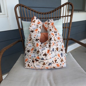 May include: A beige tote bag with a cat print design. The bag has a pocket on the front and is made of a soft, durable fabric.