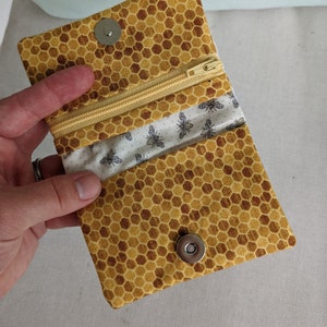 以下が含まれることがあります： 黄色いハニカム模様の小さなオープンウォレット。ファスナーとマグネットスナップ留めが付いています。内側には黒い蜂の模様の白い生地が使われています。