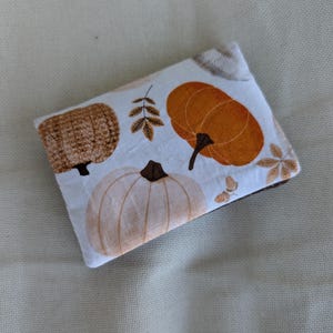 May include: A rectangular, white fabric item with a fall-themed pumpkin pattern. The design features orange, tan, and white pumpkins, along with brown leaves and stems. The item is likely a small pouch or case.