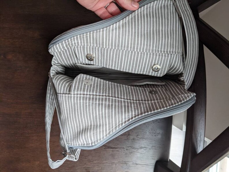 May include: A grey and white striped fabric tote bag with a zipper closure and a long strap. The bag has two pockets on the inside and a snap closure.