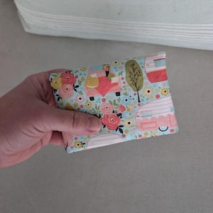 May include: A small, rectangular fabric wallet with a floral and camper design. The wallet features a light blue background with pink, yellow, and red flowers, along with illustrations of campers and trees. The wallet is held in a hand.