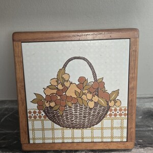 May include: A square wooden trivet with a ceramic tile insert. The tile features a brown basket overflowing with fruit, set against a white background with a dotted pattern. The border is a brown and white checkered design.