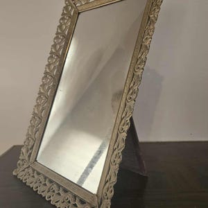 May include: A rectangular vanity mirror with an ornate, gold-colored frame featuring a detailed floral design. The mirror is angled and supported by a dark wooden stand, reflecting the surrounding environment.