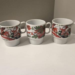 May include: Three white ceramic mugs with a festive Christmas design. Each mug features a red and green holiday pattern with a decorative rim and a curved handle. The mugs are stacked and have a ribbed base.