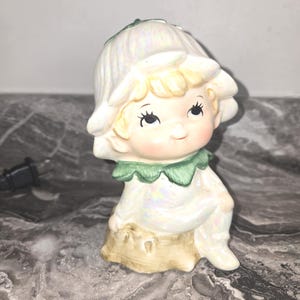 May include: A ceramic figurine of a seated child with a flower-shaped hat and a green collar. The figurine has a pearlescent finish and is sitting on a tree stump. The child has blonde hair, rosy cheeks, and is smiling.