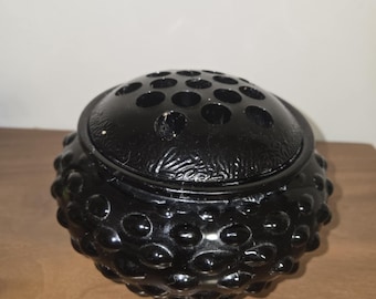 Puede incluir: Un florero de vidrio negro con un diseño de hobnail y una tapa con múltiples agujeros para arreglar los tallos. La base redonda tiene una superficie texturizada y con bultos. La tapa tiene un diseño floral decorativo.