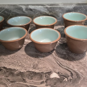 May include: A collection of eight small, round ramekins. Each ramekin has a light brown exterior, a pale blue interior, and a brown rim. The ramekins are arranged on a marbled surface.