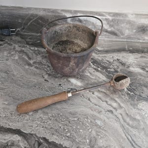 May include: A small, weathered metal bucket with a wire handle and a long-handled ladle with a wooden grip. The ladle has a small, rounded bowl at the end. Both items show signs of age and use, with a rustic, antique appearance.