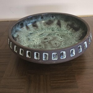 May include: A brown ceramic bowl with a unique design. The interior has a mottled green and brown glaze. The exterior features a series of rectangular cutouts around the rim, adding a decorative element to the bowl.