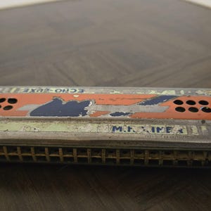 May include: Vintage ECHO-LUXE harmonica with a distressed finish. The harmonica features a silver metal body with orange and blue accents. The text "M.HOHNER" is visible on the front. A classic musical instrument.