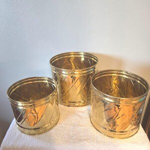 May include: Three gold-colored metal planters of varying sizes. Each planter has a cylindrical shape with a diagonal etched pattern. The planters are arranged on a white surface, reflecting light and adding a touch of elegance.