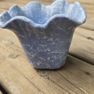 May include: A light blue ceramic bowl with a ruffled, wavy rim and a white speckled, splattered paint pattern across the exterior surface. The bowl has a wide, open top that tapers down to a smaller, rounded base, featuring a textured finish.