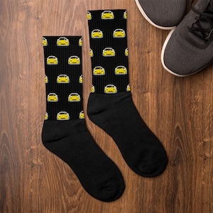 Könnte beinhalten: Schwarze Socken mit einem sich wiederholenden Muster aus gelben Sportwagen. Die Socken werden auf einer Holzoberfläche neben einem Paar grauer Turnschuhe gezeigt. Das Autodesign hat eine gelbe Karosserie mit weißen Umrissen.